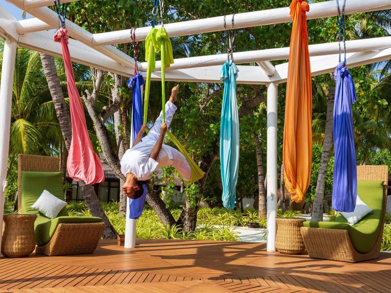 Persona practica yoga en hamacas de colores bajo una estructura de madera al aire libre con fondo verde.