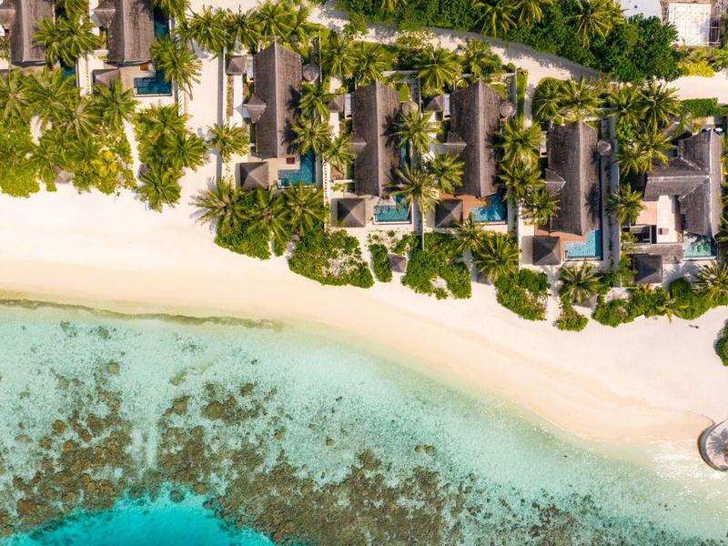 Vista aérea de bungalows en playa de arena blanca con mar turquesa y arrecifes de coral.