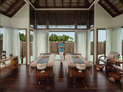 Innenraum eines Spa mit zwei Massageliegen, großen Fenstern und Blick auf grüne Pflanzen.