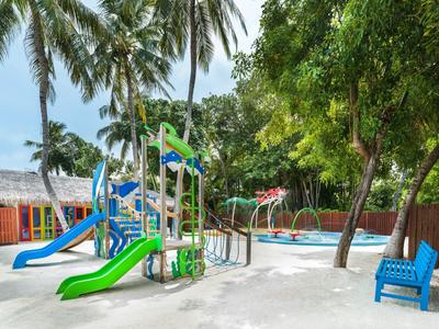 Kinderspielplatz mit Klettergerüst und Rutschen im Sand unter Palmen und Bäumen.