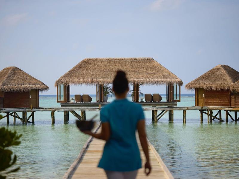 Person geht über einen Steg zu mehreren Bungalows über dem Wasser bei klarem Himmel.