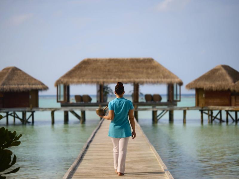Person geht auf einem Steg zu über Wasser gebauten Hütten in tropischem Hotelresort