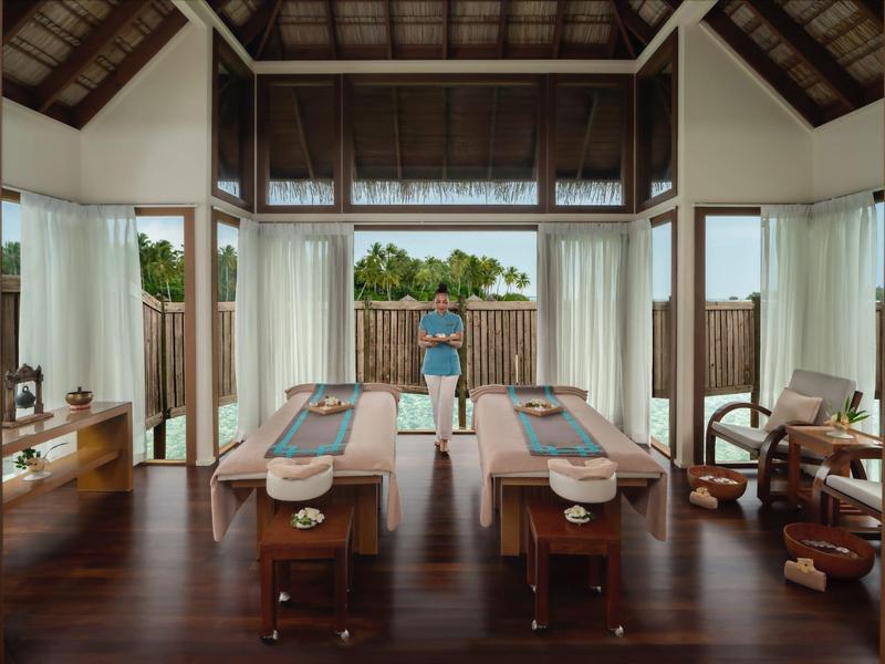 Innenraum eines Spa mit zwei Massageliegen, großen Fenstern und Blick auf grüne Pflanzen.