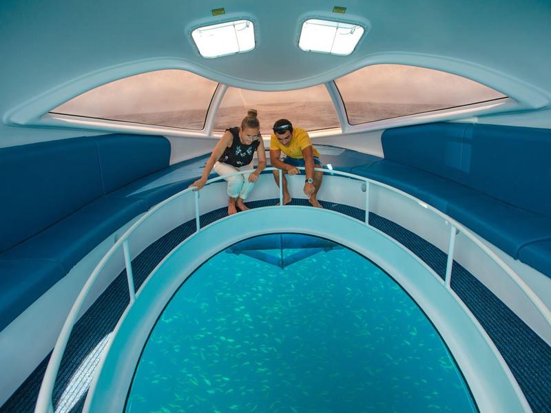 Zwei Personen sitzen in einem Boot mit einem gläsernen Boden und blicken auf das Wasser.