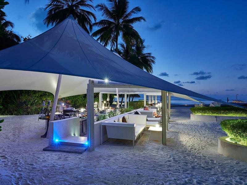 Abendliche Strandlounge mit weißem Zelt, Sitzbereichen und Palmen im Hintergrund.