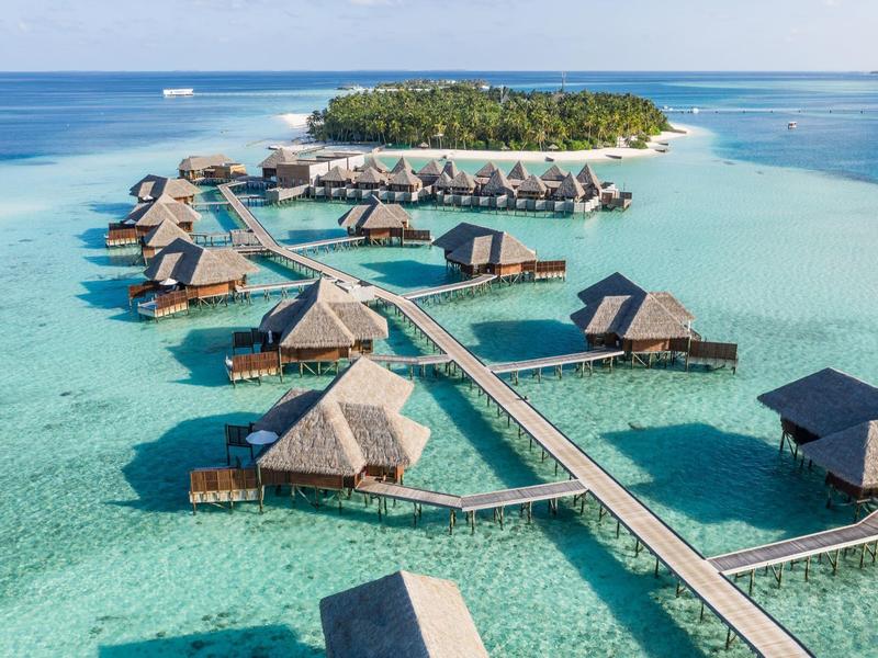 Überwasserbungalows in klarem türkisblauem Wasser mit einem bewaldeten Inselhintergrund.