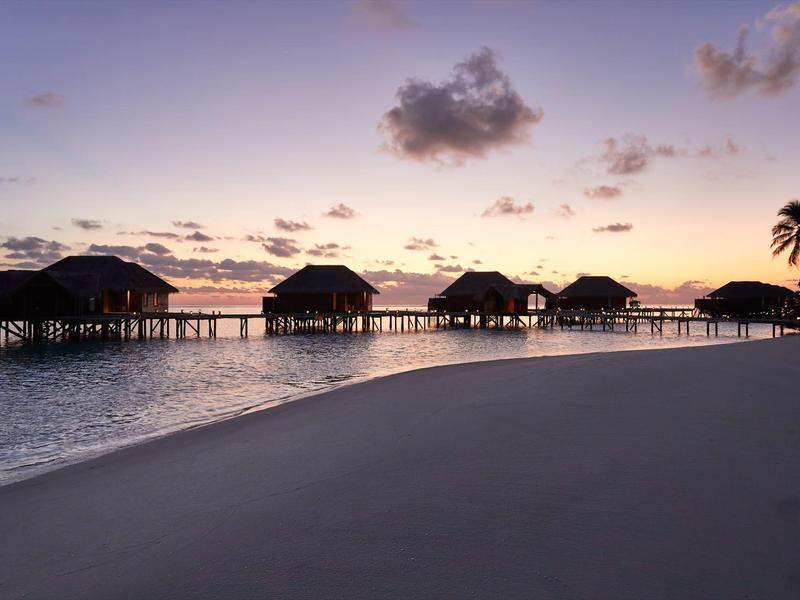 Bungalows auf Stelzen über dem Wasser bei Sonnenuntergang an einem Strand mit Palmen.