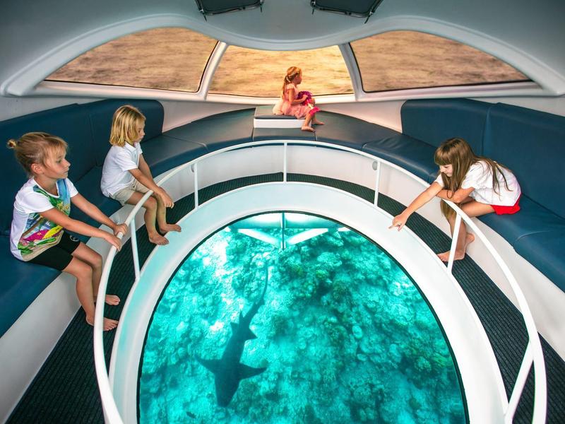 Kinder sitzen auf einem Boot mit großem Glasboden und betrachten das klare blaue Wasser darunter.