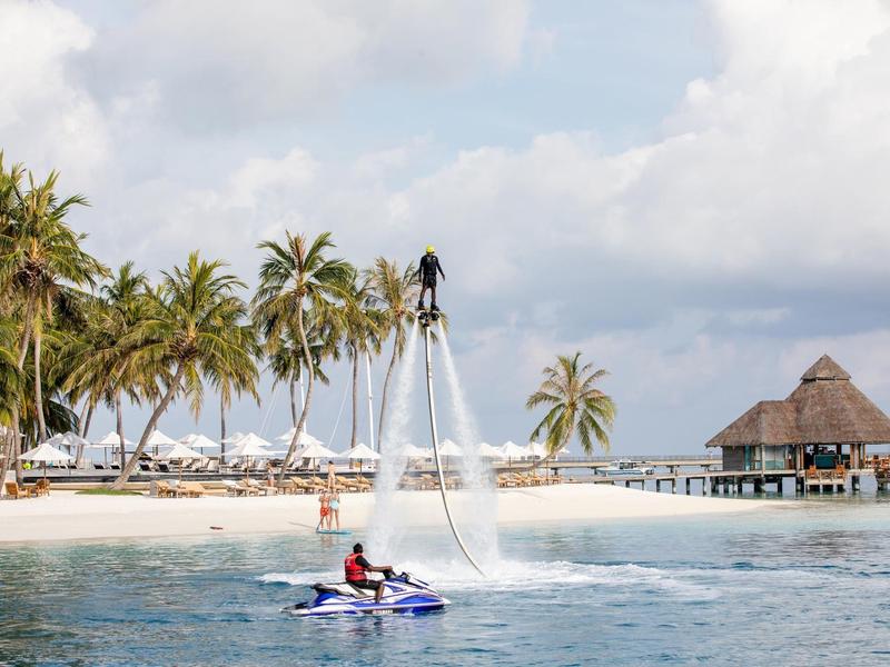 Person fliegt mit Wasser-Jetpack über Wasser vor tropischem Strand mit Palmen und Hütte.