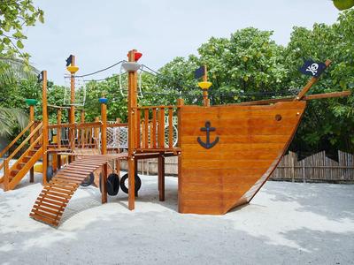 Un parco giochi a forma di nave pirata in legno sulla sabbia con alberi verdi sullo sfondo.