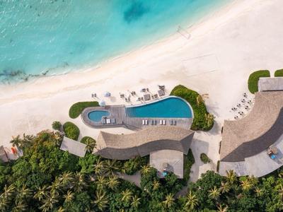 Vista aerea di una piscina accanto a una spiaggia di sabbia bianca con ombrelloni e vegetazione tropicale.