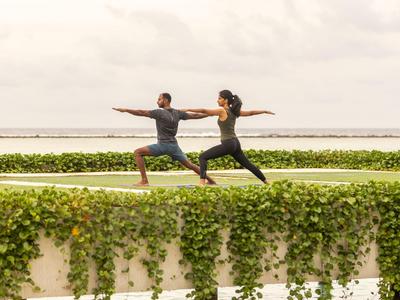 Deux personnes pratiquent la posture du guerrier en yoga sur un pont couvert de verdure au-dessus de l'eau.