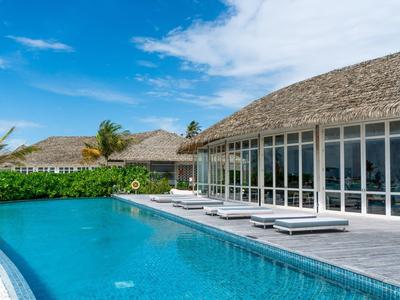Piscina moderna con sedie a sdraio davanti a un hotel con facciata in vetro e tetto di paglia sotto un cielo blu.