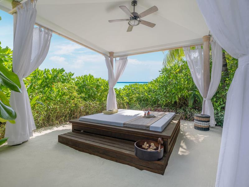 Padiglione aperto con letto e vista sul mare, circondato da piante verdi.
