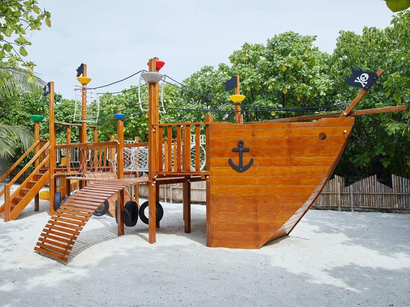 Un parco giochi a forma di nave pirata in legno sulla sabbia con alberi verdi sullo sfondo.