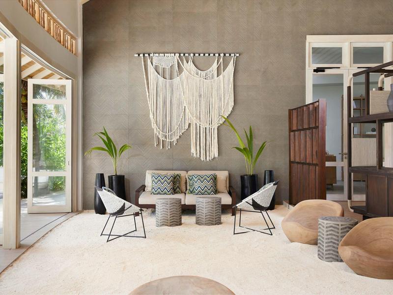 Area lounge moderna con appeso murale in macramé, poltrone e palette di colori neutri.