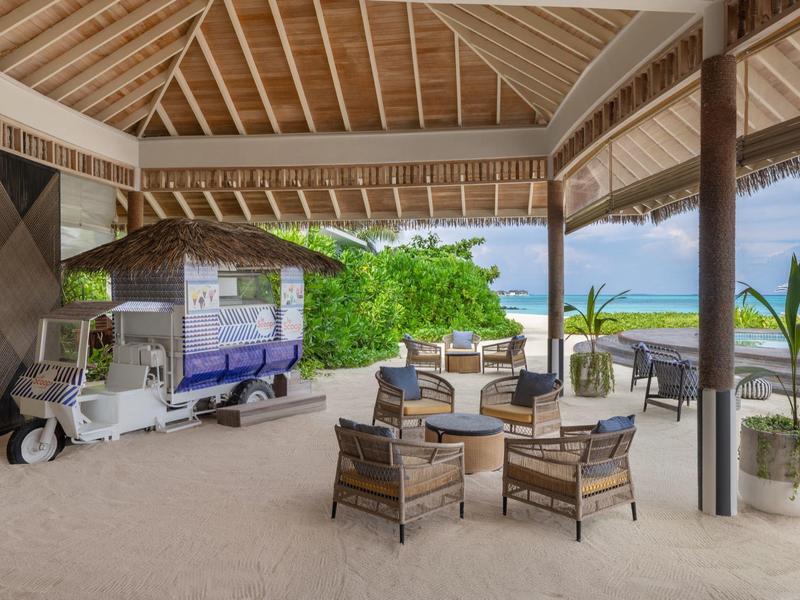 Padiglione aperto sulla spiaggia con sedie e tavoli, vista sul mare e piante verdi.