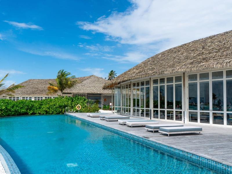 Piscina moderna con sedie a sdraio davanti a un hotel con facciata in vetro e tetto di paglia sotto un cielo blu.
