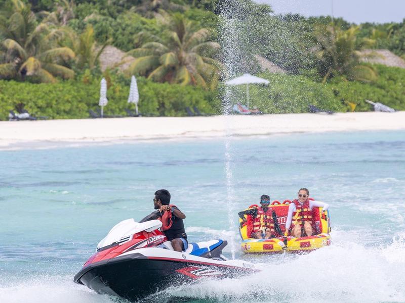 Persona su moto d'acqua che traina diverse persone su un tubo giallo gonfiabile nel mare vicino a una spiaggia.