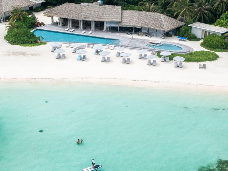 Resort sulla spiaggia con sabbia bianca, piscina e ospiti in acqua turchese.
