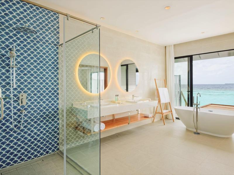 Helles Badezimmer mit freistehender Badewanne, rundem Spiegel und blau gekachelter Dusche mit Glaswand.