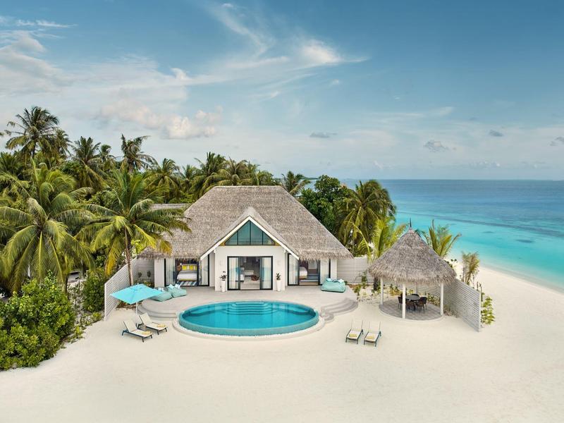 Luxuriöse Strandvilla mit runder Pool, Palmen, weißen Sand und blauem Meer im Hintergrund.