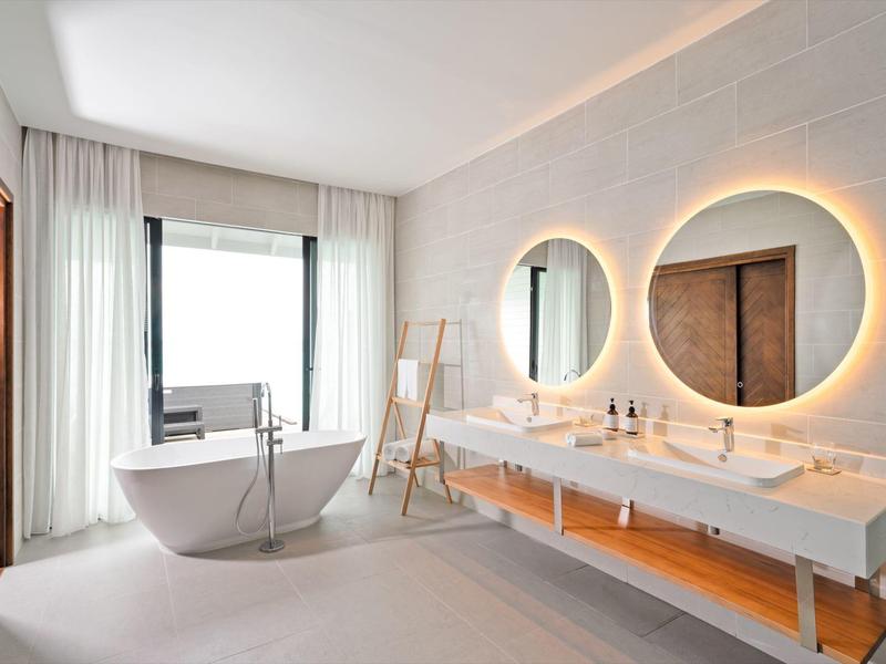 Helles Bad mit freistehender Badewanne, zwei runden beleuchteten Spiegeln und weißen Waschtischen.