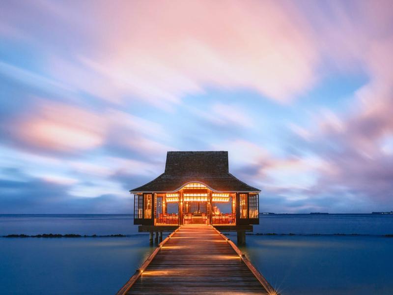 Ein von Laternen beleuchteter Holzsteg führt zu einem Häuschen über ruhigem Wasser bei buntem Abendhimmel.