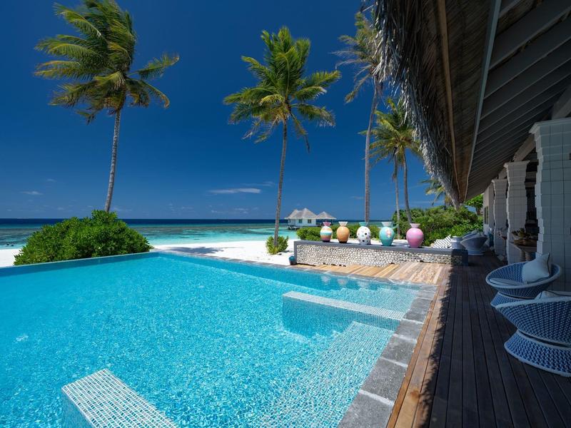 Piscina moderna y zona de salón junto a la playa con palmeras bajo el cielo azul.
