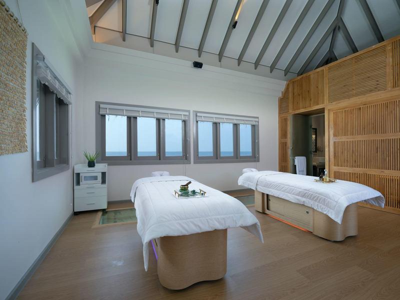 Sala de tratamientos luminosa con dos camillas de masaje, ventana amplia y pared revestida en madera.