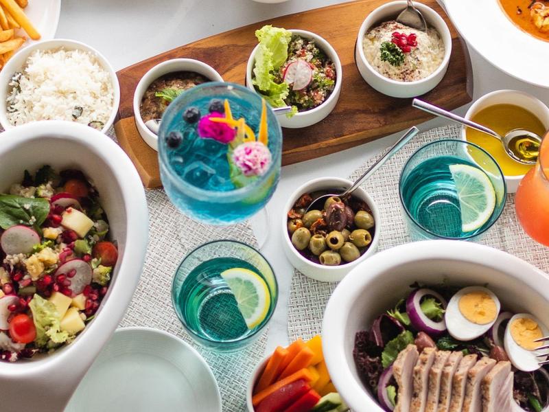 Mesa con variedad de platos coloridos, ensaladas, bebidas y aperitivos, ambiente de comida gourmet.