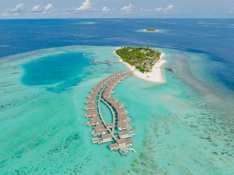 Vista aérea de un resort en una pequeña isla con bungalows sobre el agua en océano turquesa.
