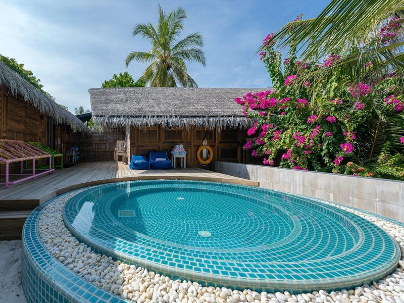 Un jacuzzi redondo al aire libre frente a una cabaña con plantas florecientes y palmeras de fondo.