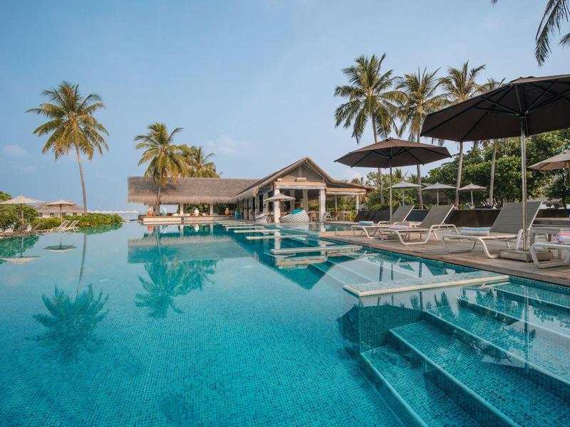 Piscina infinita junto a un restaurante en un resort tropical con palmeras y tumbonas.