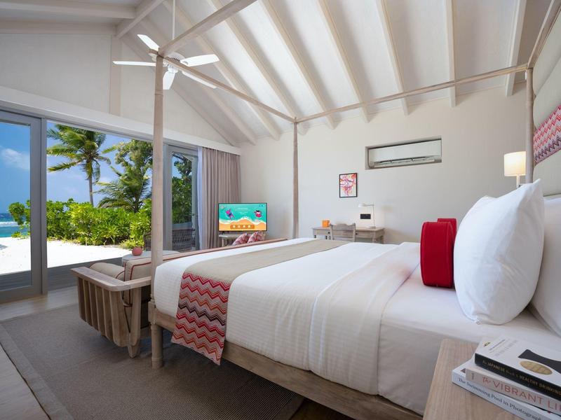 Habitación de hotel moderna con cama grande, ventilador de techo y vista a jardín tropical y mar.