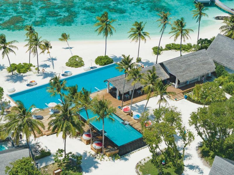 Vista aérea de una playa tropical con piscina, palmeras y pabellones abiertos en un resort.