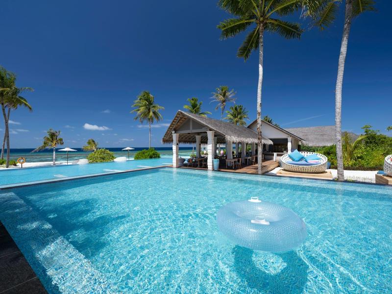 Piscina clara con palmeras y un pabellón en un resort tropical bajo cielo azul.