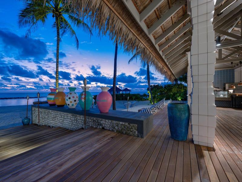 Vista nocturna de una terraza de madera con vistas al mar y palmeras en un destino vacacional.