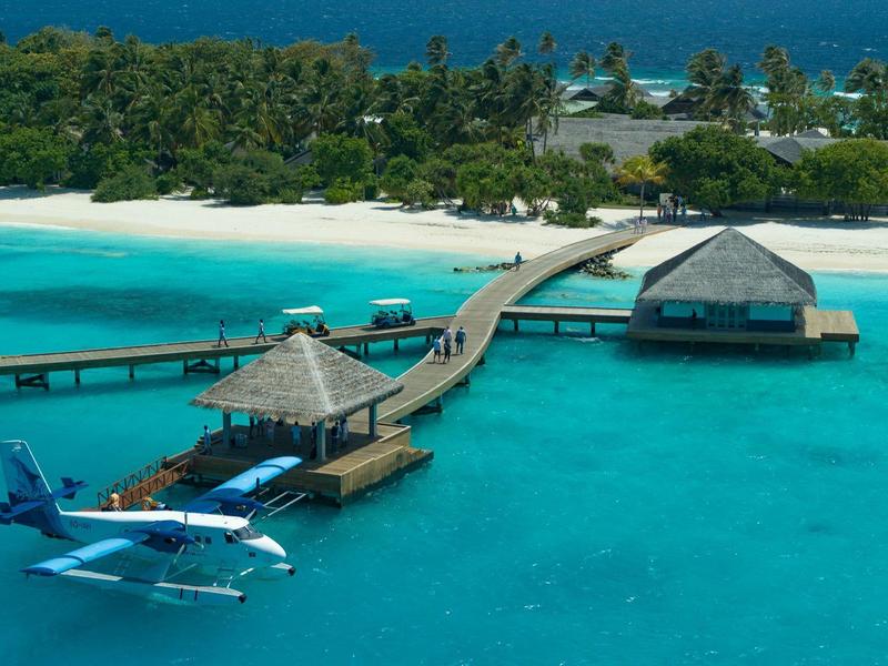 Vista aérea de una isla tropical con bungalows sobre el agua y un hidroavión en el muelle.
