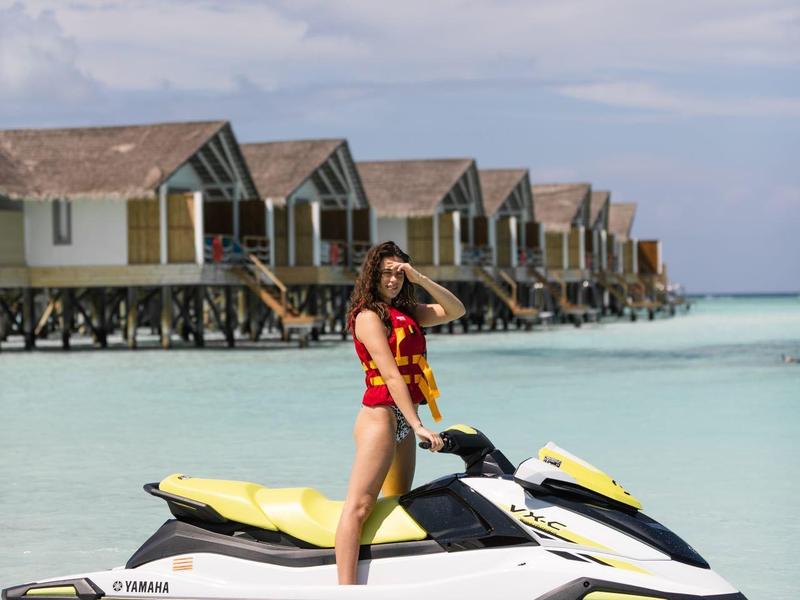 Mujer en moto acuática en agua clara frente a una fila de bungalows sobre pilotes.