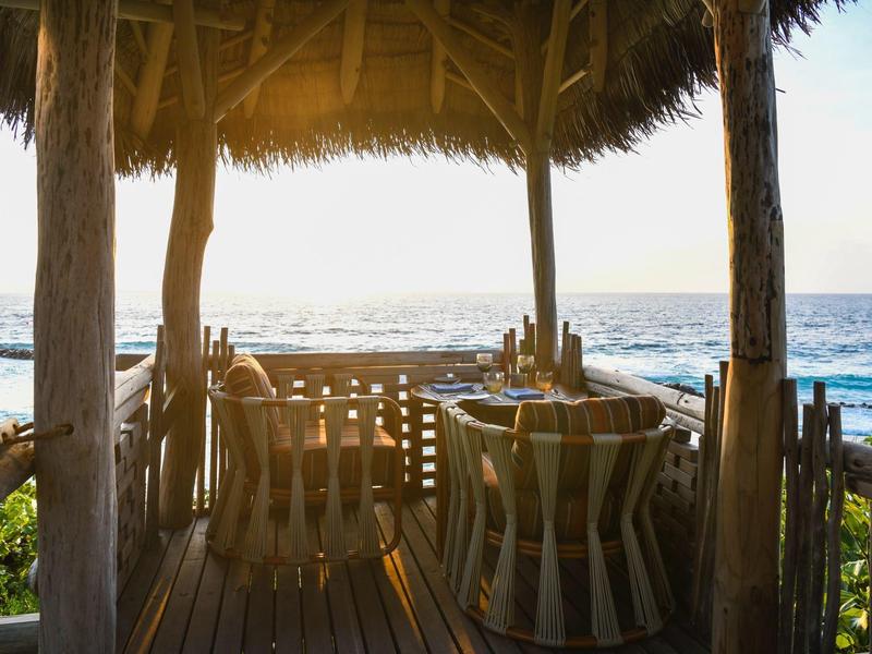 Terraza cubierta con sillas y mesas con vista al mar al atardecer.