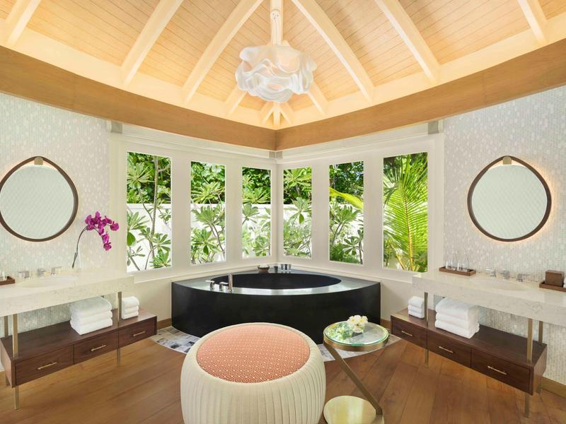 Baño lujoso con bañera negra, grandes ventanas, espejos y taburete en tonos cálidos.