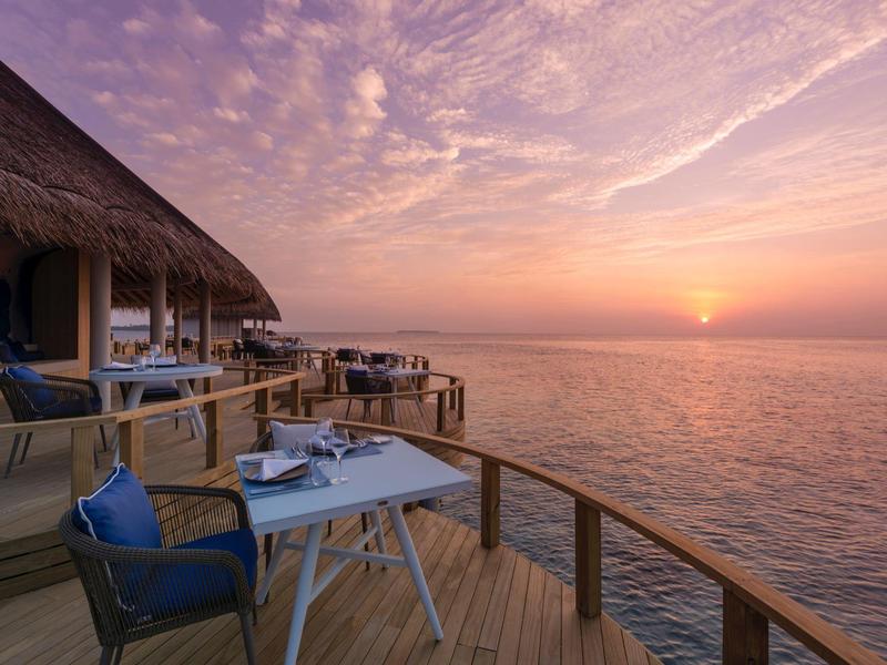 Vue depuis une terrasse en bois avec des chaises et des tables sur la mer au coucher du soleil.