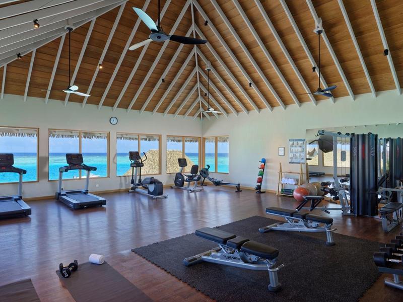Salle de sport lumineuse avec grandes fenêtres, plafond en bois et vue sur la mer.