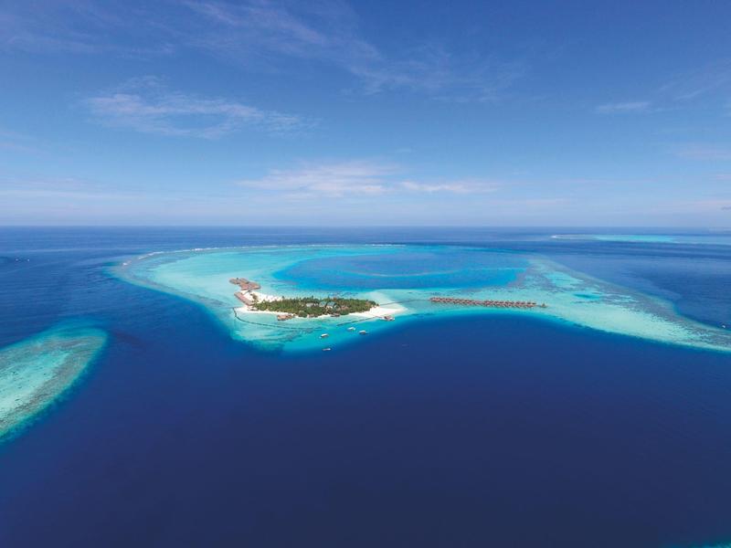 A small tropical island with a surrounding coral reef in clear blue ocean under a partly cloudy sky.