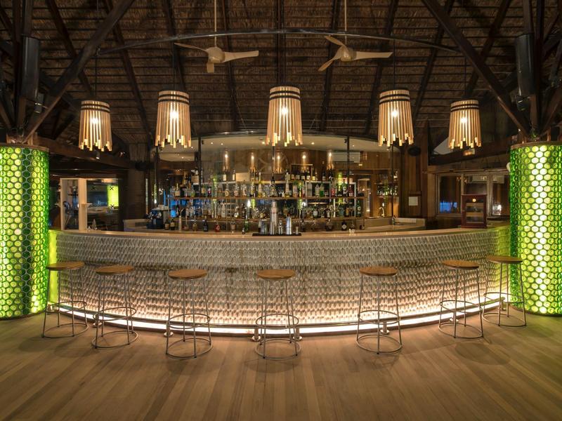 Modern hotel bar with lit counter, metal stools, and green-lit pillars under wooden roof.