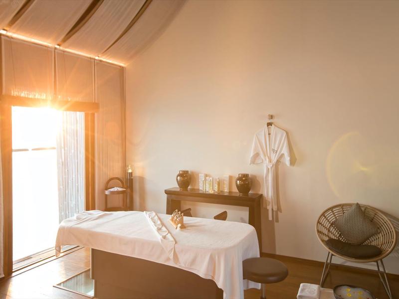 Hotel room with sunlit bed, wooden furniture, and cozy chair near a balcony.