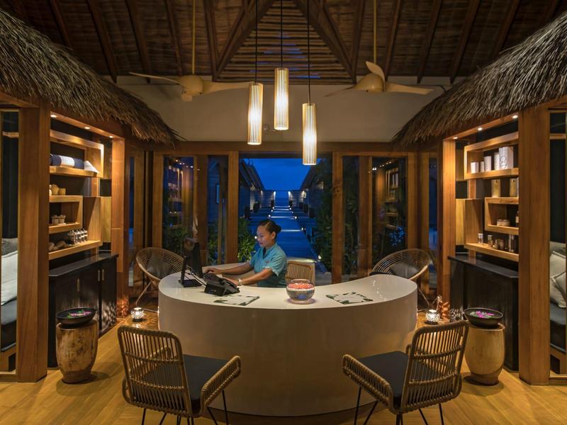 Cozy tropical reception area with wooden furniture, thatched roof, and a staff member at the desk.