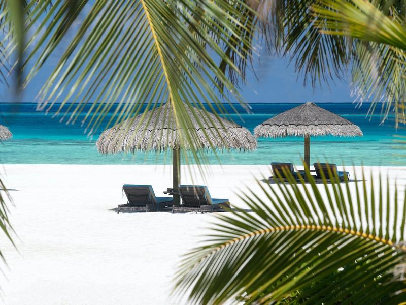 White sandy beach with palm trees, two lounge chairs, and straw umbrellas by clear blue sea.