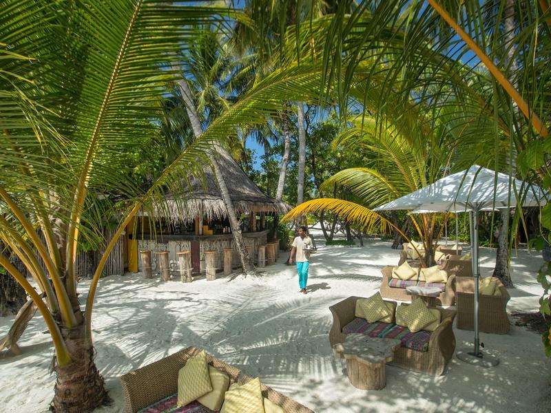 Beachfront seating area with umbrellas and palm trees surrounding sandy ground.